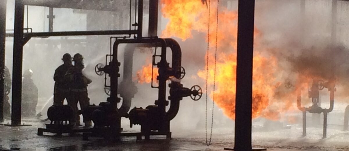 Firefighters operating near burning industrial piping during a fire response.