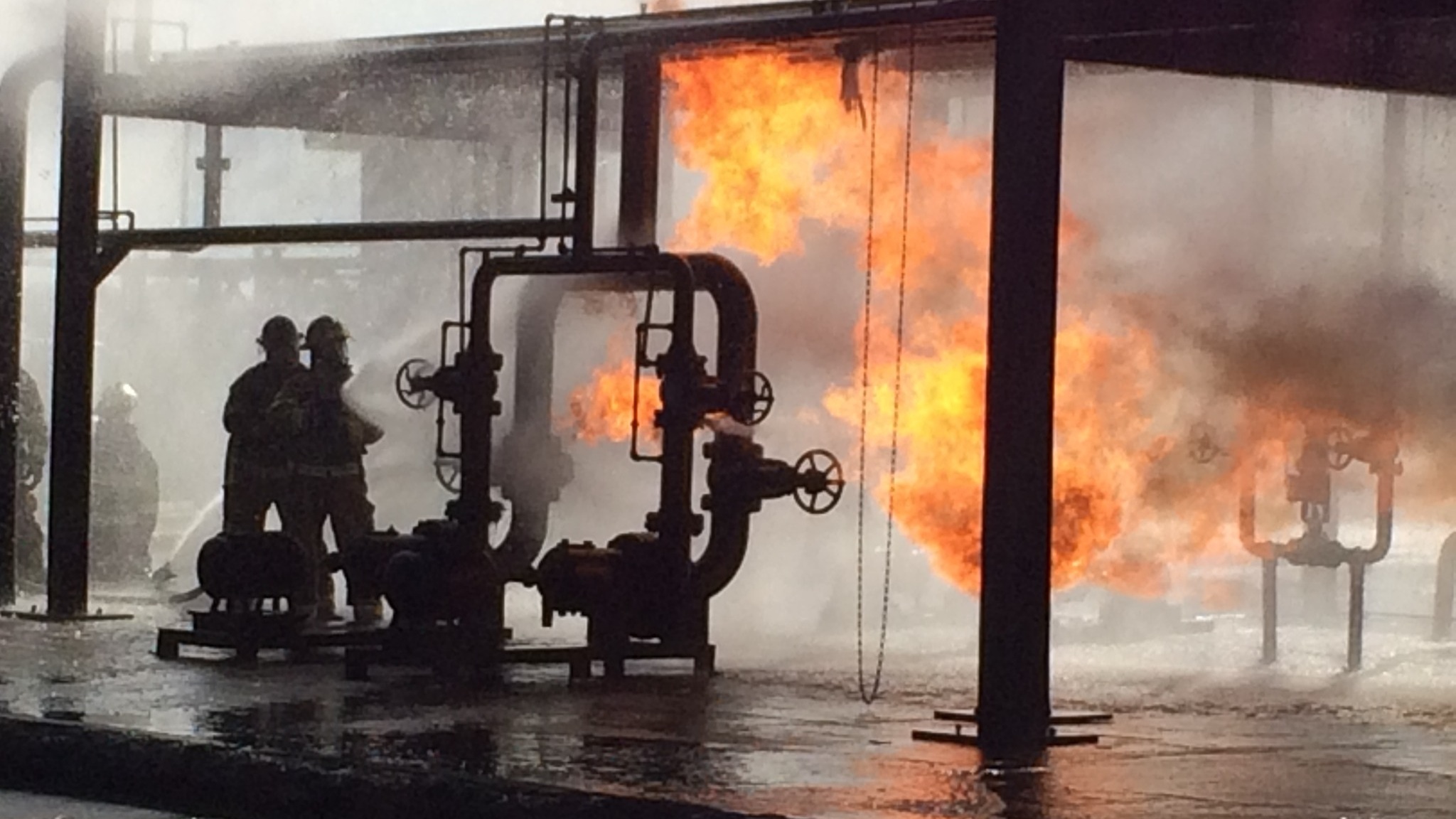 Firefighters operating near burning industrial piping during a fire response.